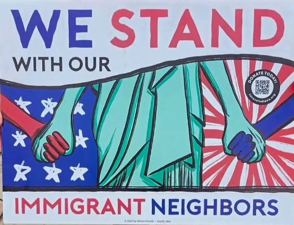 A yard sign that says We stand with our immigrant neighbors. The Statue of Liberty holds hands with a person on each side, over a flag of stars on one side and red and white stripes on the other.